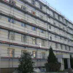 Wielopiętrowy budynek w trakcie prac elewacyjnych, otoczony rusztowaniem na tle błękitnego nieba.